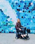 Amy, a disabled, white Australian woman with blonde hair, sits on her mobility scooter in front of a wall with blue and white geometric shapes. 