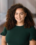 Carmen, a biracial Black woman with curly hair, wearing a dark green dress and a pearl necklace, smiles warmly against a blurred background.