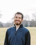 The image is a headshot of Kevin Andrews in a grassy field smiling. A young, Caucasian male, he has brown hair, a beard, and is wearing a color-blocked quarter zip sweater with a blue body and black sleeves. There is a shimmer of light from the sun illuminating his face. In the blurred background you can see the grassy field with buildings and scattered bare trees.